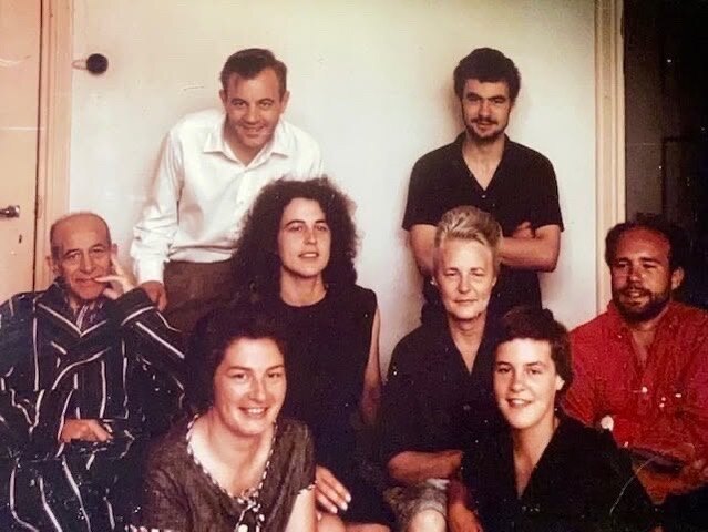 Groepsfoto van acht mensen van verschillende leeftijden, binnen voor een witte muur