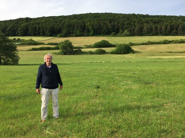 Roel van Duijn staat in een groot grasveld voor een beboste heuvel